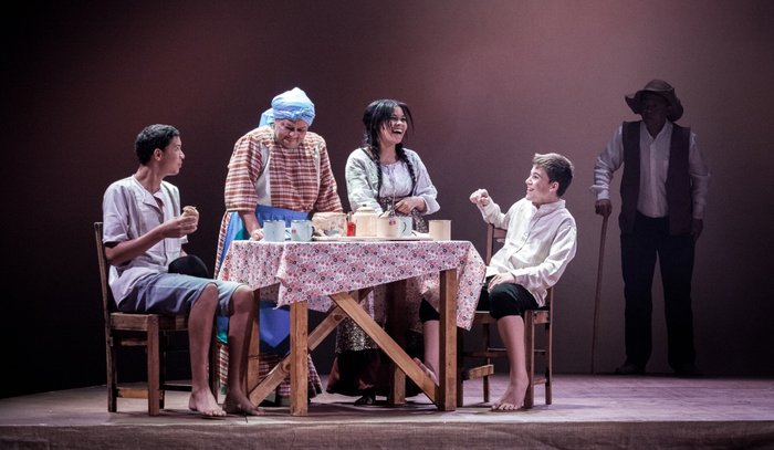 Vyf karakters om 'n tafel met 'n geblomde tafeldoek; 'n man met 'n hoed staan eenkant in die skaduwee.